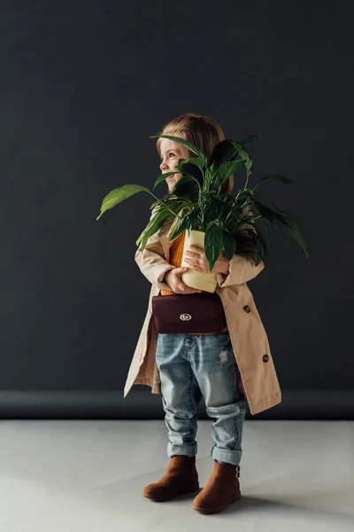 Mignon enfant en trench coat et jeans tenant la plante en pot de fleurs sur fond noir — Photo de stock