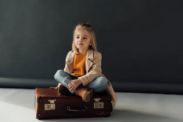 Задумчивый ребенок сидит со скрещенными ногами на кожаном чемодане на черном фоне — стоковое фото