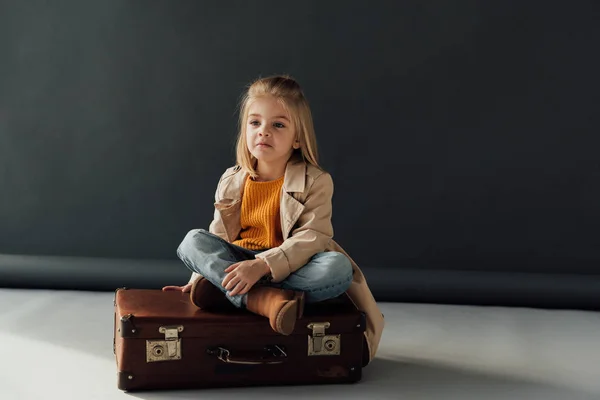 Nachdenkliches Kind sitzt mit gekreuzten Beinen auf Lederkoffer auf schwarzem Hintergrund — Stockfoto