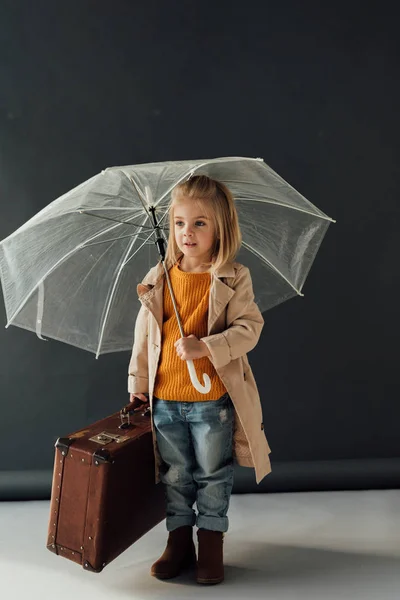 Enfant en trench coat et jeans tenant parapluie et valise en cuir sur fond noir — Photo de stock