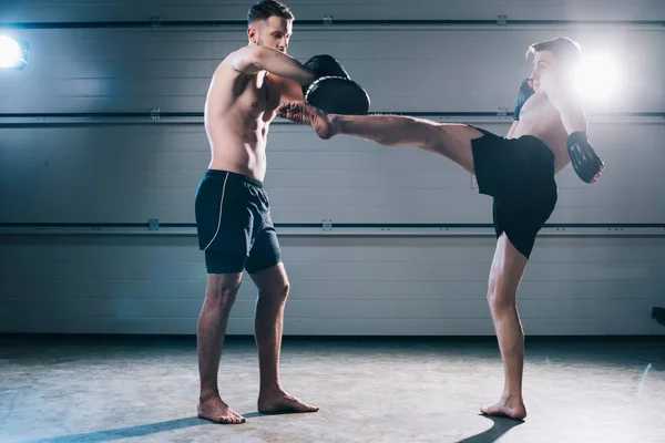 Fort musclé torse nu mma combattant pratiquant coup de pied élevé avec un autre sportif pendant l'entraînement — Photo de stock