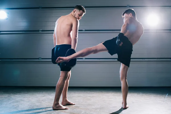 Back view of strong muscular barefoot mma fighter practicing low kick with another sportsman — Stock Photo