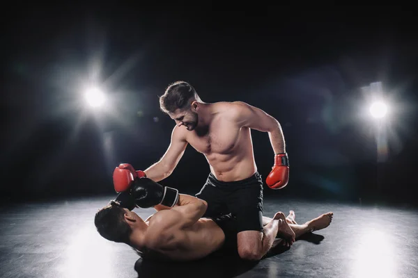 Strong mma fighter in boxing gloves punching opponent while sportsman lying on floor — Stock Photo
