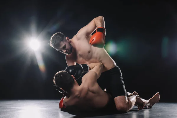 Shirtless strong mma fighter in boxing gloves punching opponent in head while sportsman lying on floor — Stock Photo