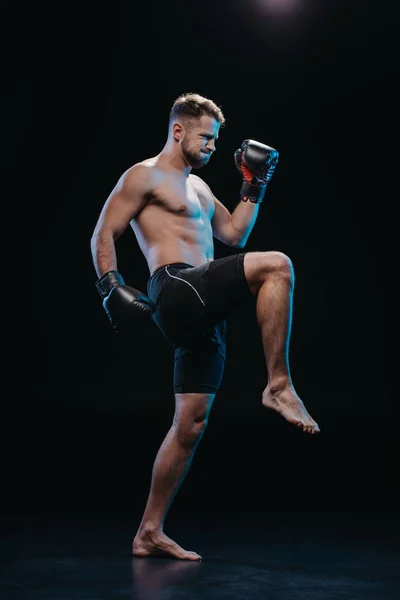 Muscular barefoot strenuous boxer in boxing gloves doing kick on black — Stock Photo