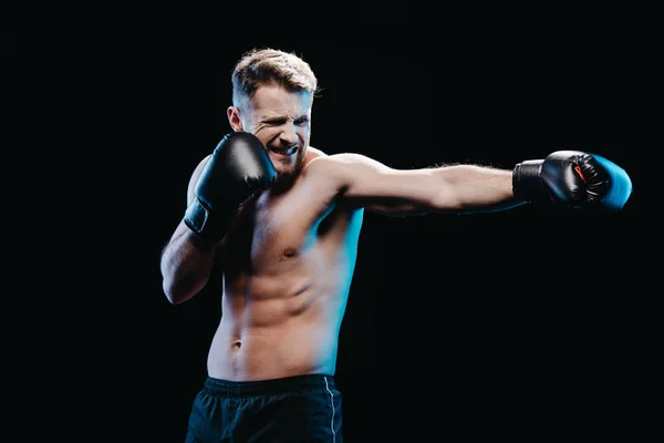 Muscular sporty boxer with strenuous face expression in boxing gloves doing punch isolated on black — Stock Photo