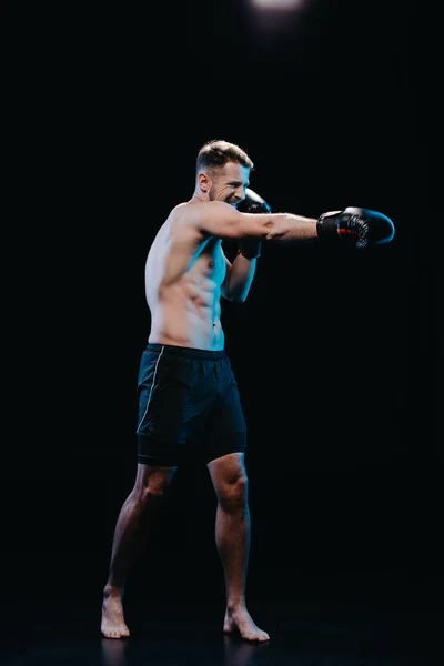 Barefoot sporty boxer with strenuous face expression in boxing gloves doing punch isolated on black — Stock Photo