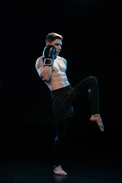 Muscular barefoot mma fighter in boxing gloves doing kick on black — Stock Photo