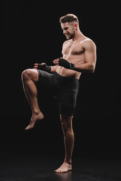 Muscular barefoot mma fighter in bandages doing kick with knee on black — Stock Photo