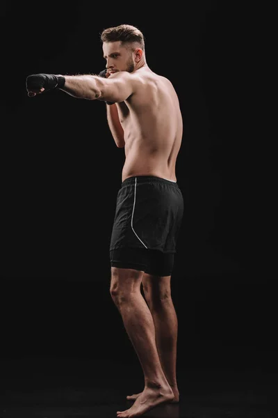 Сильный мускулистый боец Мма в бинтах и шортах, делающий пунш на черном — стоковое фото