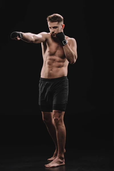 Strong barefoot muscular mma fighter in bandages and shorts doing punch on black — Stock Photo