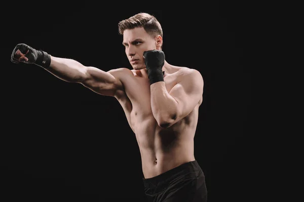 Strong shirtless muscular sportsman in bandages doing punch isolated on black — Stock Photo
