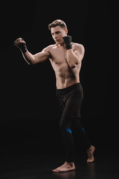 Strong barefoot muscular sportsman in bandages doing punch on black — Stock Photo