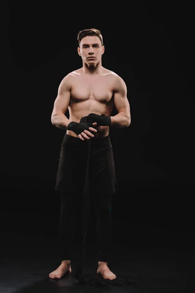 Strong shirtless and barefoot muscular mma fighter fixing bandages and looking at camera on black — Stock Photo