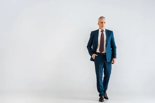 Pensive mature businessman in suit standing with hand in pocket on grey — Stock Photo