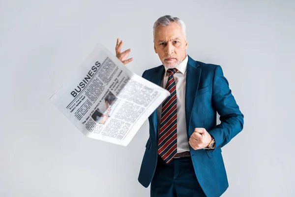 Angry mature man in formal wear throwing business newspaper while standing isolated on grey — Stock Photo