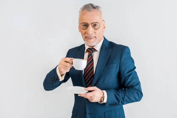 Cheerful mature businessman in formal wear holding saucer and cup isolated on grey — Stock Photo