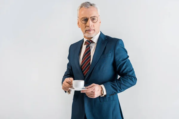 Ernsthafte reife Geschäftsmann in formeller Kleidung mit Untertasse und Tasse isoliert auf grau — Stockfoto
