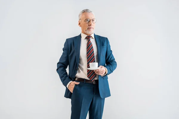 Serious mature businessman holding saucer with cup and standing with hand in pocket isolated on grey — Stock Photo