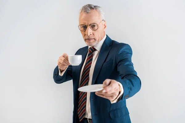 Selbstbewusster, reifer Geschäftsmann in formeller Kleidung, Untertasse und Tasse isoliert auf grau haltend — Stockfoto
