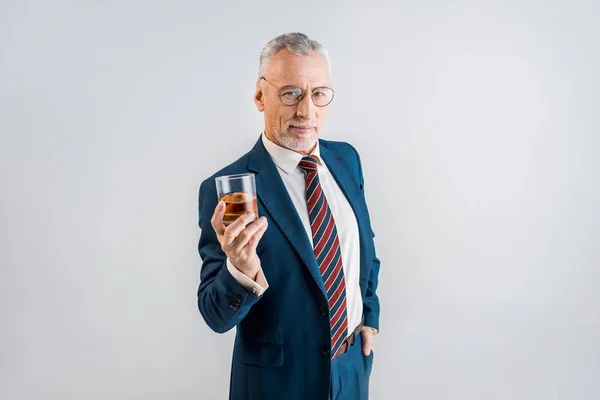 Alegre hombre de negocios maduro sosteniendo vaso de whisky mientras está de pie con la mano en el bolsillo aislado en gris - foto de stock