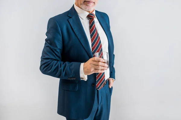 Cropped view of mature businessman holding glass of whiskey while standing with hand in pocket isolated on grey — Stock Photo