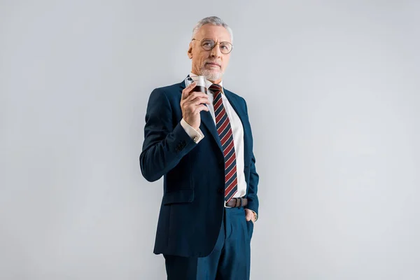 Reifer Geschäftsmann mit einem Glas Whiskey im Stehen mit der Hand in der Tasche, isoliert auf grau — Stockfoto