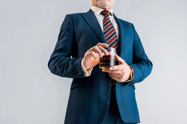 Vista recortada de hombre de negocios maduro en traje sosteniendo vidrio con whisky aislado en gris - foto de stock