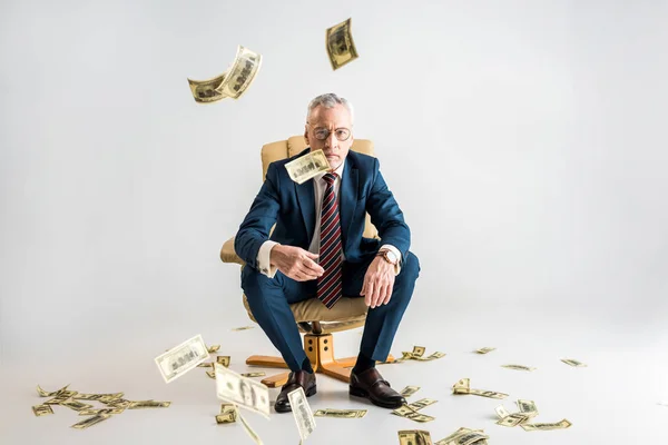 Selective focus of mature businessman sitting in armchair and throwing in air dollar banknotes on grey — Stock Photo