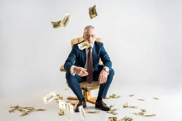 Enfoque selectivo de hombre de negocios maduro sentado en sillón y lanzando billetes de banco en dólares de aire en gris - foto de stock