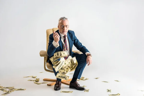 Selective focus of serious mature businessman sitting in armchair and throwing in air dollar banknotes on grey — Stock Photo