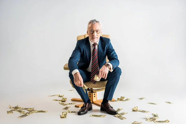 Homme d'affaires mature joyeux assis dans un fauteuil et tenant des billets en dollars sur gris — Photo de stock