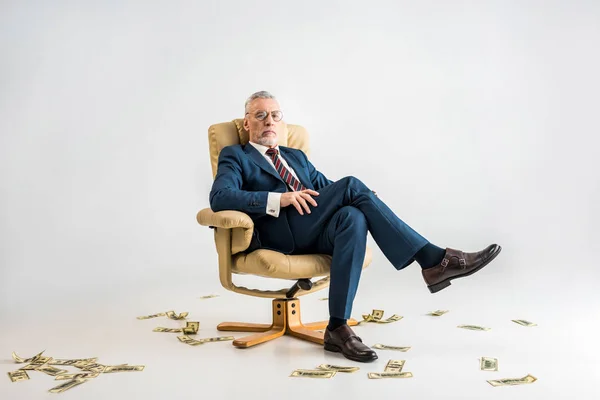 Confident mature businessman sitting in armchair near dollar banknotes on grey — Stock Photo
