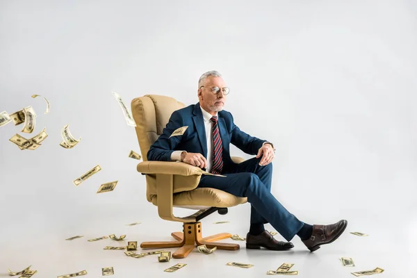 Selektiver Fokus eines selbstbewussten reifen Geschäftsmannes, der im Sessel neben Dollarnoten auf grau sitzt — Stockfoto
