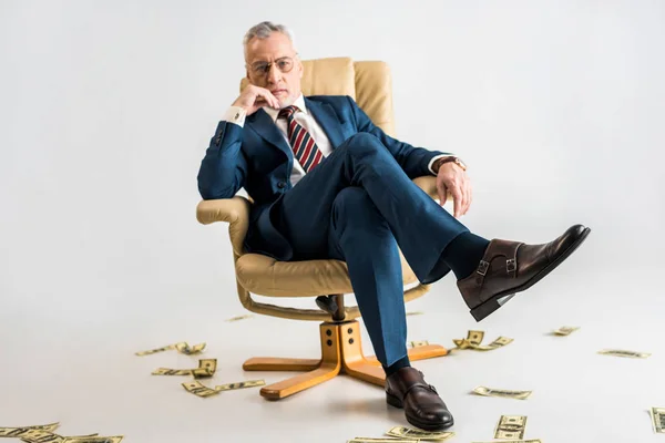 Pensive mature businessman sitting in armchair near dollar banknotes on grey — Stock Photo