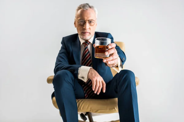 Селективное внимание зрелого бизнесмена, сидящего в кресле и держащего стакан виски, изолированного на сером — стоковое фото