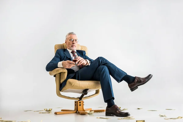 Mature businessman in suit sitting in armchair near dollar banknotes and holding glass of whiskey on grey — Stock Photo