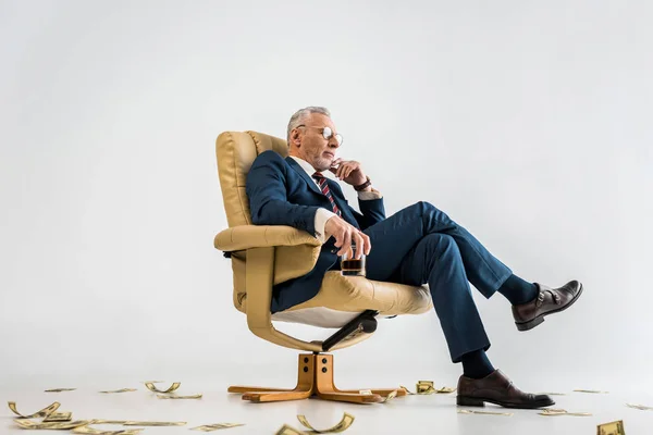 Pensativo hombre de negocios maduro sentado en sillón cerca de billetes de dólar y sosteniendo vaso de whisky en gris - foto de stock