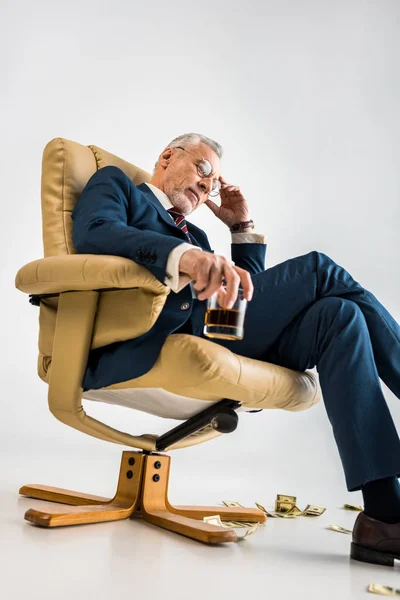 Vue à angle bas de coûteux homme d'affaires mature assis dans un fauteuil près de billets en dollars et tenant un verre de whisky sur gris — Photo de stock