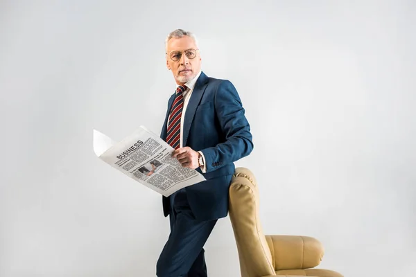 Homem de negócios maduro de pé perto de poltrona e segurando jornal de negócios isolado em cinza — Fotografia de Stock