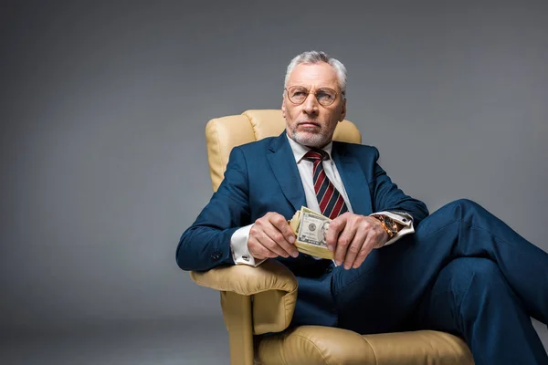 Pensive mature businessman sitting in armchair and holding dollar banknotes on grey — Stock Photo