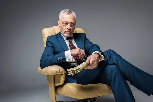 Selektiver Fokus eines reifen Geschäftsmannes, der im Sessel sitzt und Dollarnoten in grau hält — Stockfoto