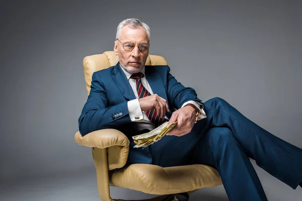 Selective focus of mature businessman sitting in armchair and holding dollar banknotes on grey — Stock Photo