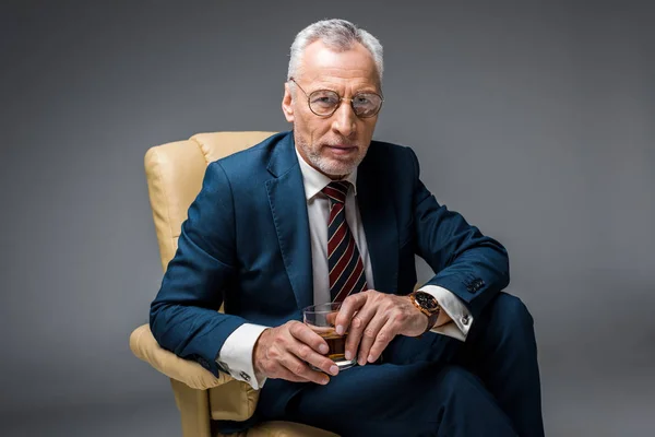 Mature businessman sitting in armchair and holding glass of whiskey on grey — Stock Photo