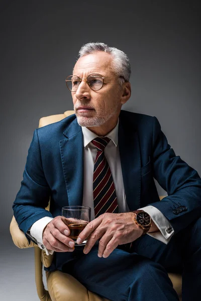 Reifer Geschäftsmann sitzt im Sessel und hält ein Glas Whiskey auf grau — Stockfoto