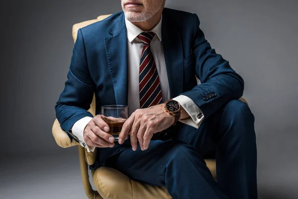 Vista recortada de hombre de negocios maduro en traje sentado en sillón y sosteniendo vaso de whisky en gris - foto de stock