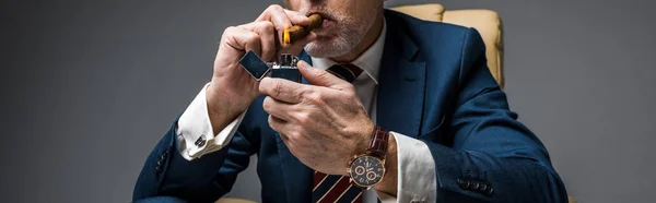 Panoramic shot of mature businessman holding lighter while smoking cigar on grey — Stock Photo