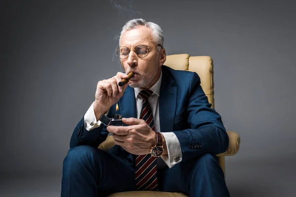 Mature man in suit holding lighter while smoking cigar with closed eyes on grey — Stock Photo