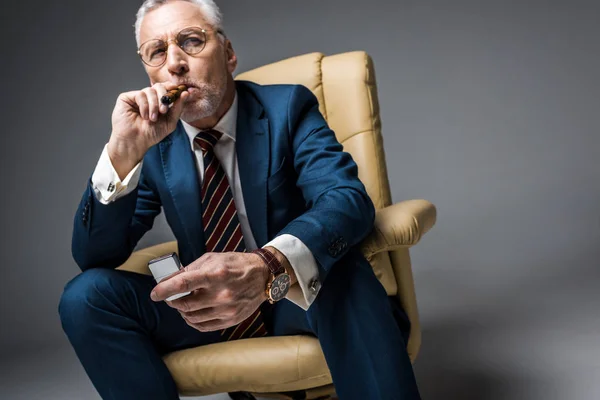 Hombre maduro en gafas y traje sosteniendo más ligero mientras fuma cigarro en gris - foto de stock