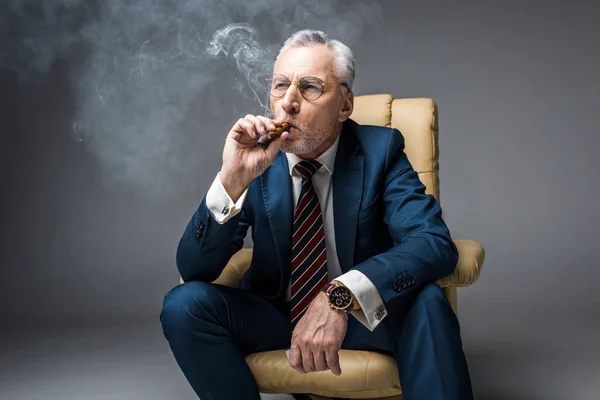 Homem maduro em óculos e terno fumar charuto enquanto sentado em poltrona no cinza — Fotografia de Stock